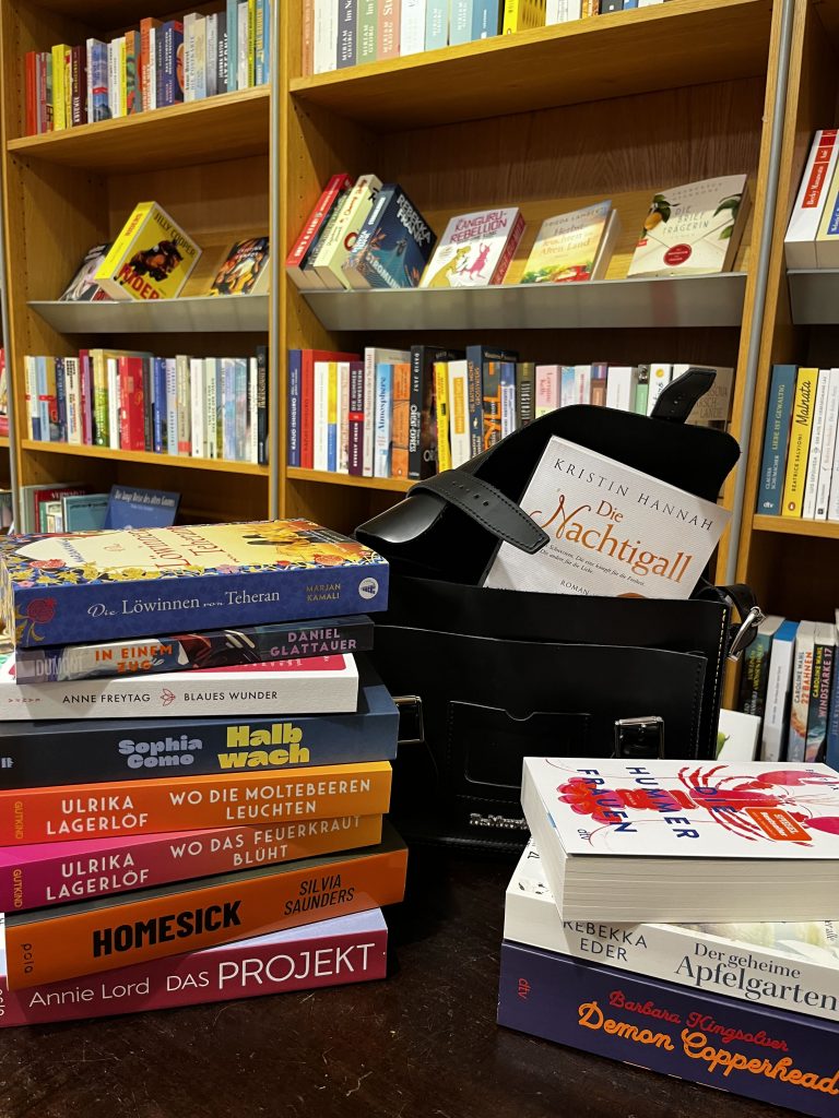 Eine Foto mit Bücherregalen im Hintergrund. Im Vordergrund liegen auf einem dunklen Holztisch zwei Stapel Taschenbücher und eine offene Handtasche, aus der ebenfalls ein Taschenbuch herausguckt.