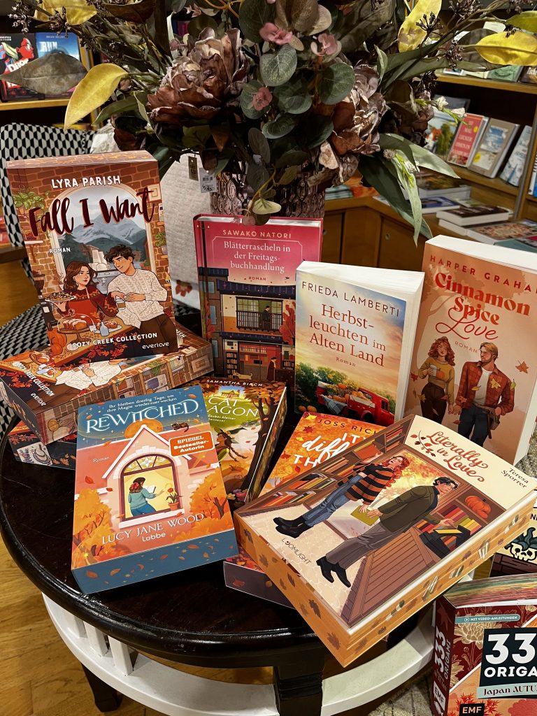 Auf einem runden, dunklen Holztisch steht ein großer Strauß aus Plastikblumen. Drumherum sind verschiedene herbstlich gestaltete Taschenbücher und Hardcover Bücher angeordnet.