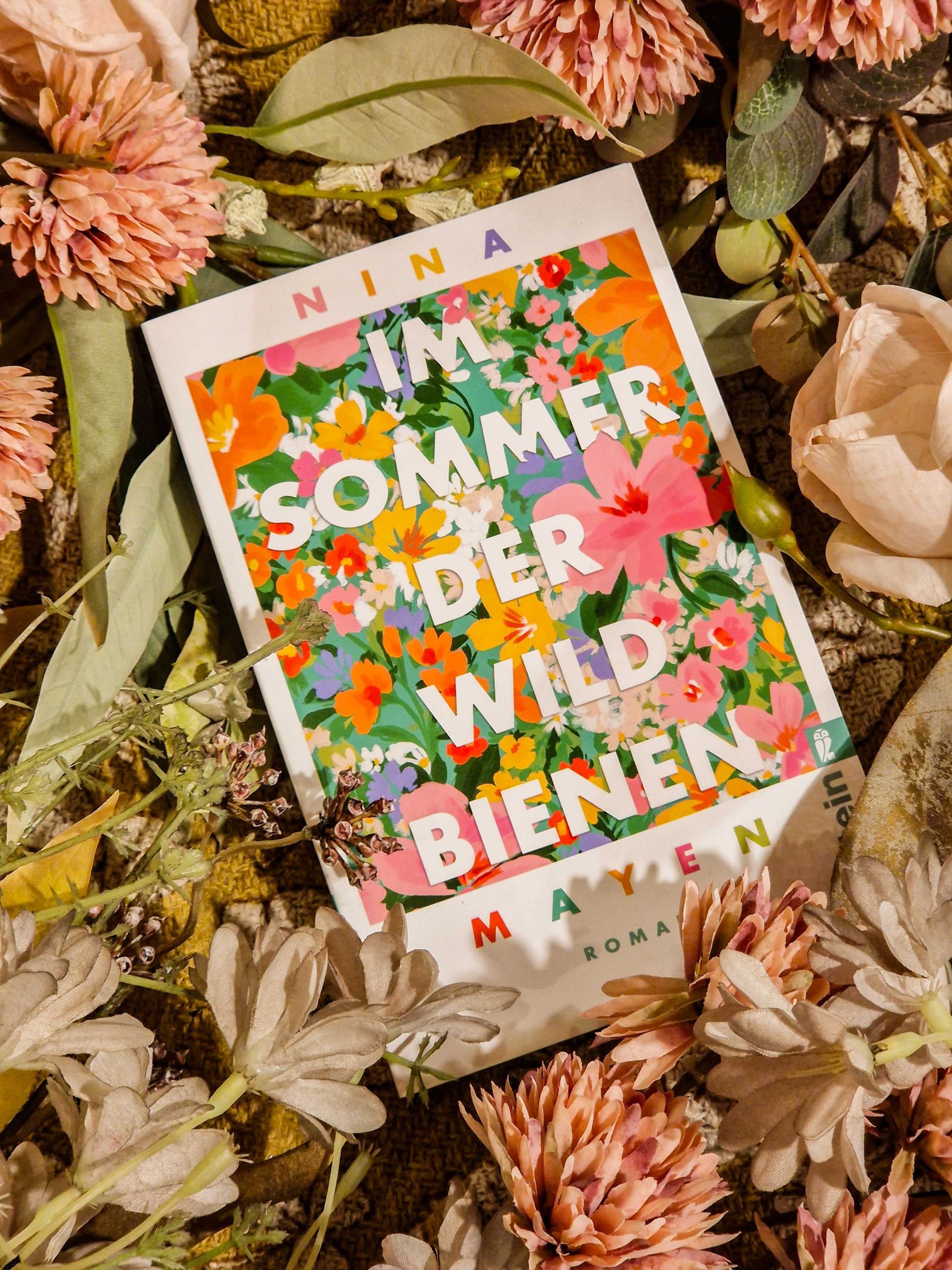 Ein Foto mit dem Cover von "Im Sommer der Wildbienen" umgeben von einer Menge Kunstblumen und Pflanzen