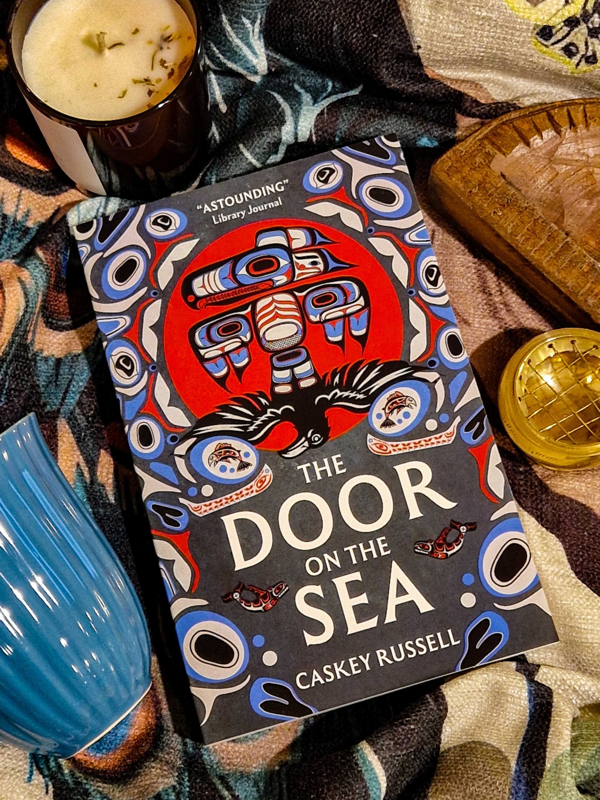 Ein Foto von dem Buch "The Door on the Sea" auf einer bunten Decke, umgeben von einer Kerze, einer blauen Tasse und kleineren Dekoartikeln