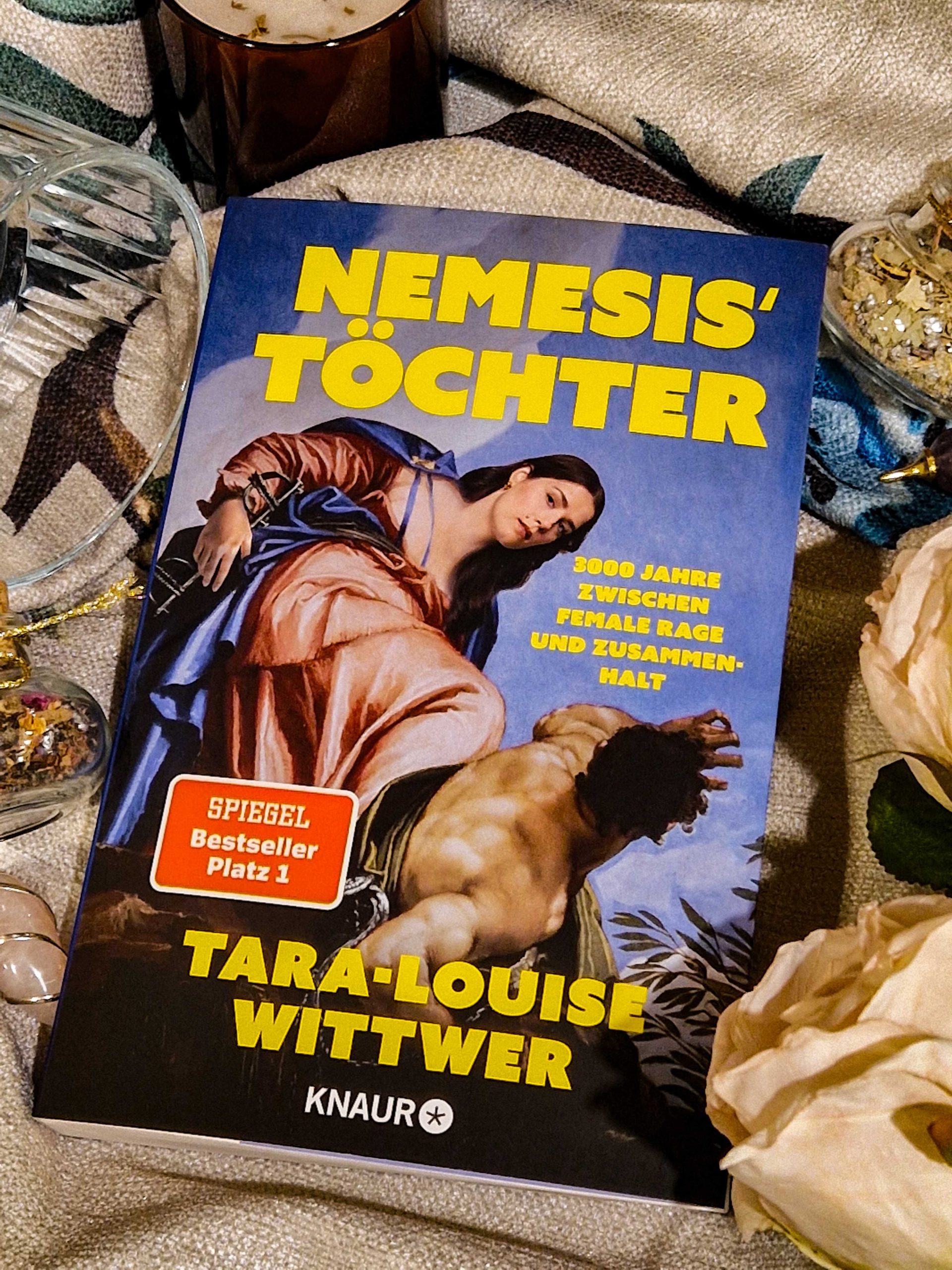 Ein Foto mit dem Cover von "Nemesis' Töchter" umgeben von verschiedenen Dekoartikeln